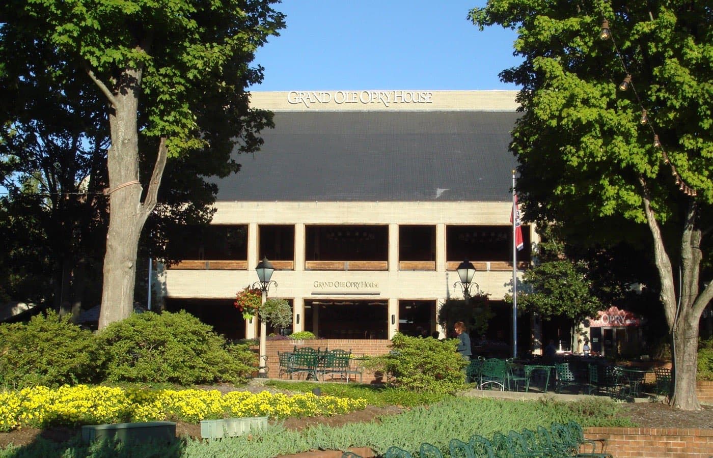 The front of the Grand Ole Opry House on a sunny day, with a “Grand Ole Opry House” sign on the roofline and over the entrance, framed by two large trees above a garden of yellow flowers and green café chairs