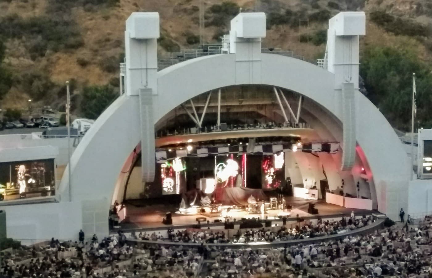 The Hollywood Bowl's white arched shell at dusk, a lit stage and video screens under it, with a full crowd in the seats and a scrubby hillside behind