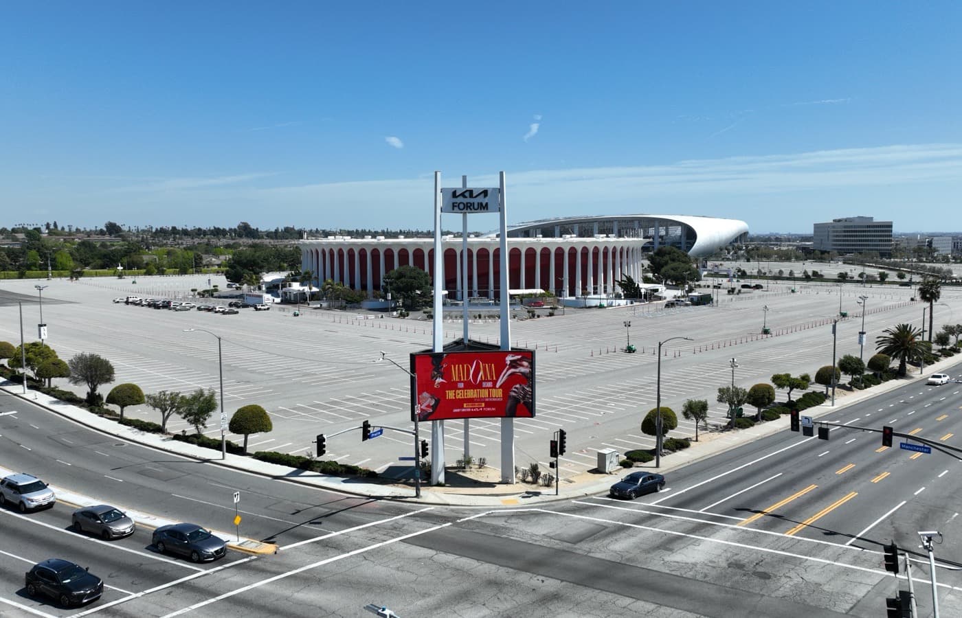 The Kia Forum's round white colonnade with red panels behind a wide, empty parking lot, with a tall Kia Forum sign and a lit billboard at the corner of Prairie Avenue and Manchester Boulevard in front, and a large curved white roof rising behind the Forum
