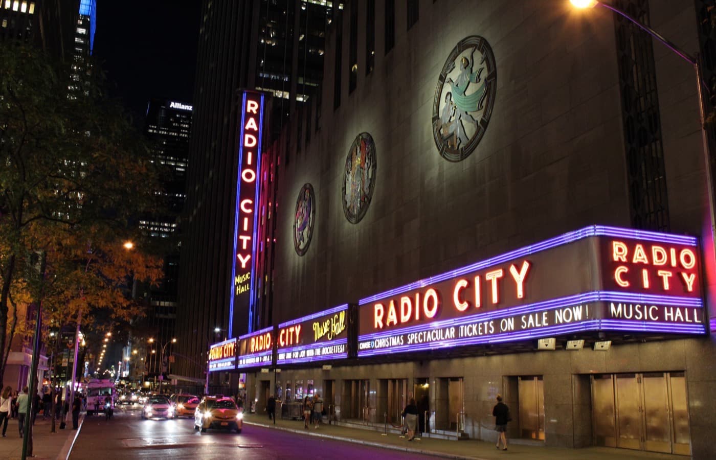 Radio City Music Hall at night, its tall vertical Radio City Music Hall sign and long neon marquee lit in red and purple along the street, with round Art Deco plaques on the stone facade above, taxis on the street and people walking past the entrance doors