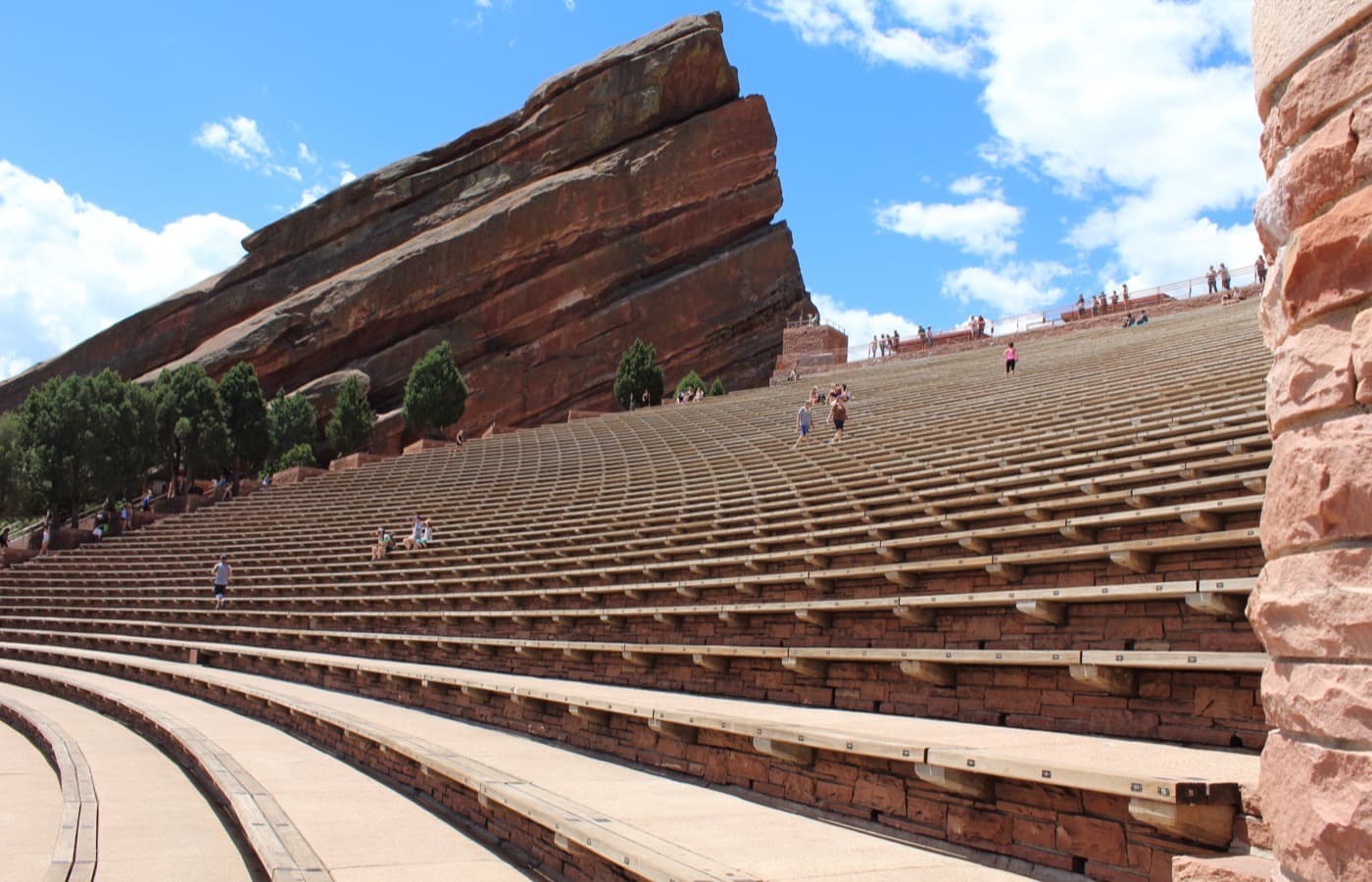 Rows of curved wooden benches climbing the red sandstone amphitheatre, with a huge tilted red rock and pine trees on the left under a blue sky