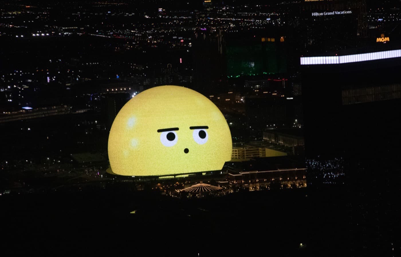 Sphere at night seen from high above, its dome lit bright yellow with a cartoon face, among dark Las Vegas buildings and city lights